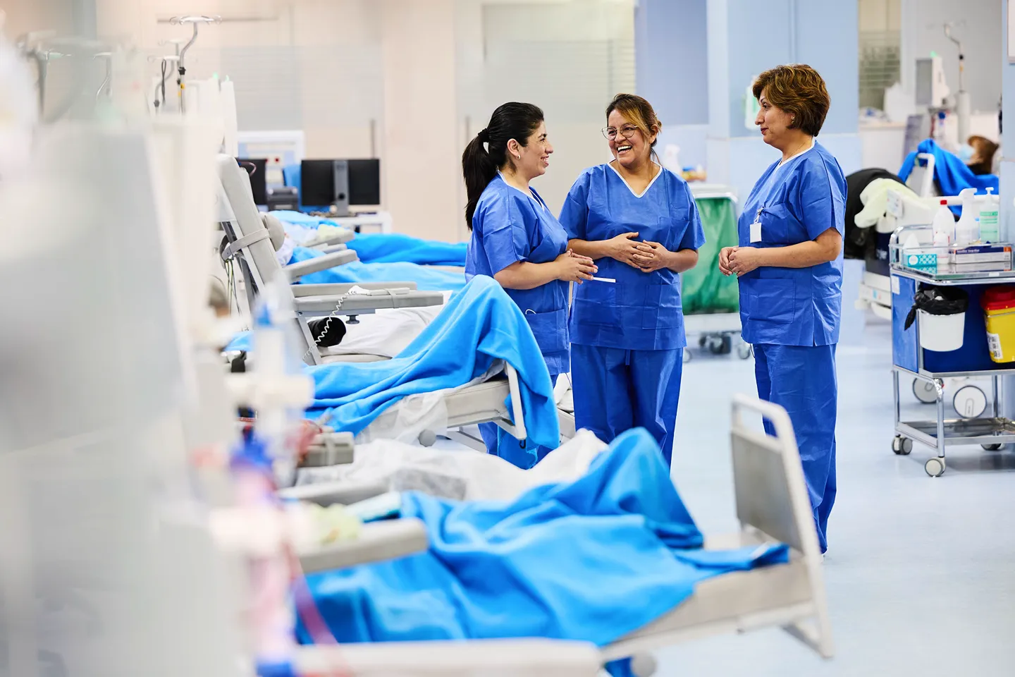 Three healthcare workers in blue scrubs stand and talk together in a brightly lit hospital room, surrounded by patient beds and medical equipment. They appear to be smiling and engaged in conversation.