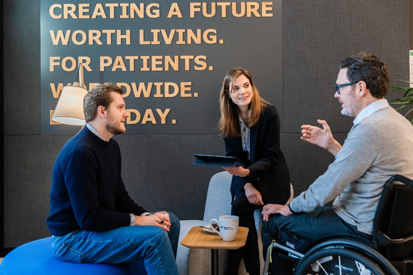 Four professionals are having a conversation in a modern office lobby. One person is in a wheelchair. The wall behind them displays the Fresenius Medical Care logo.