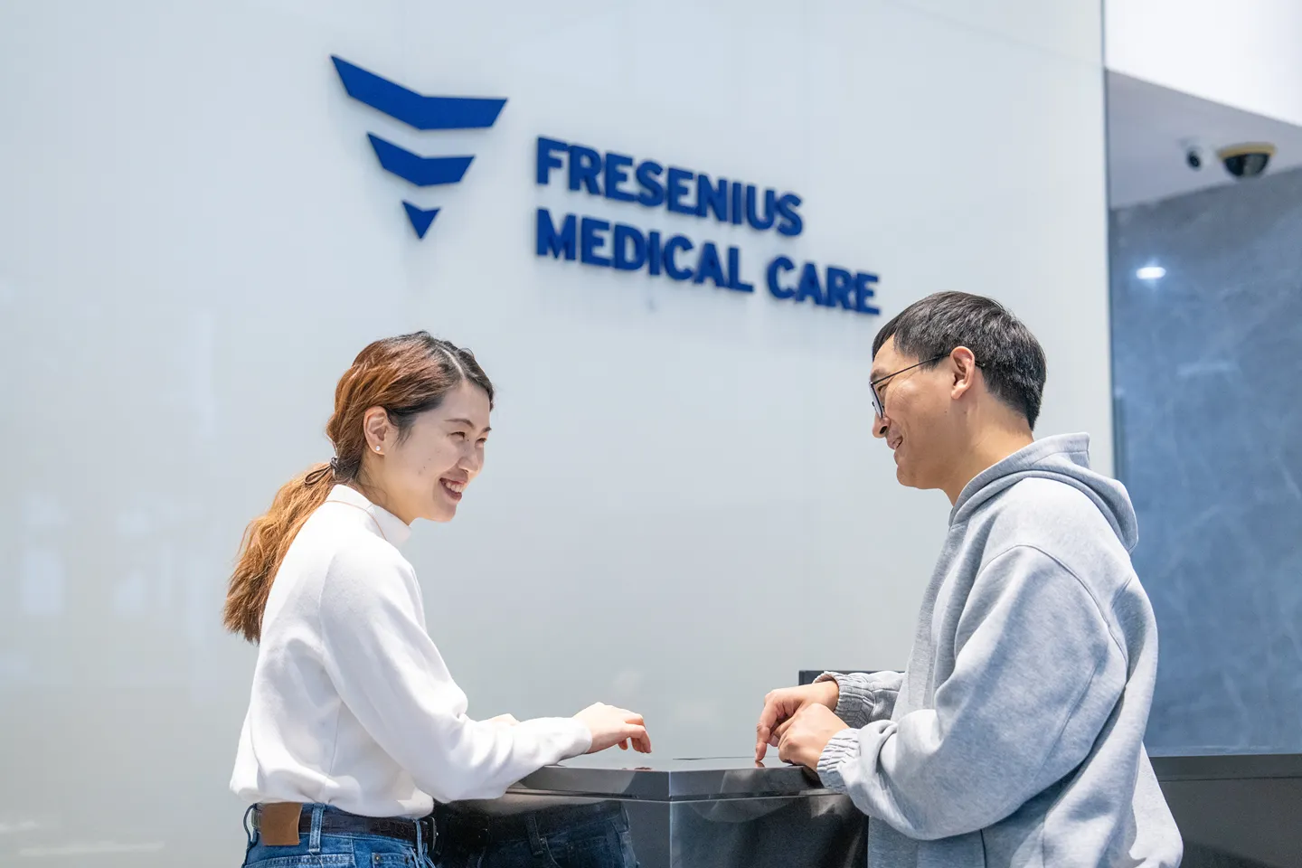 Four professionals are having a conversation in a modern office lobby. One person is in a wheelchair. The wall behind them displays the Fresenius Medical Care logo.