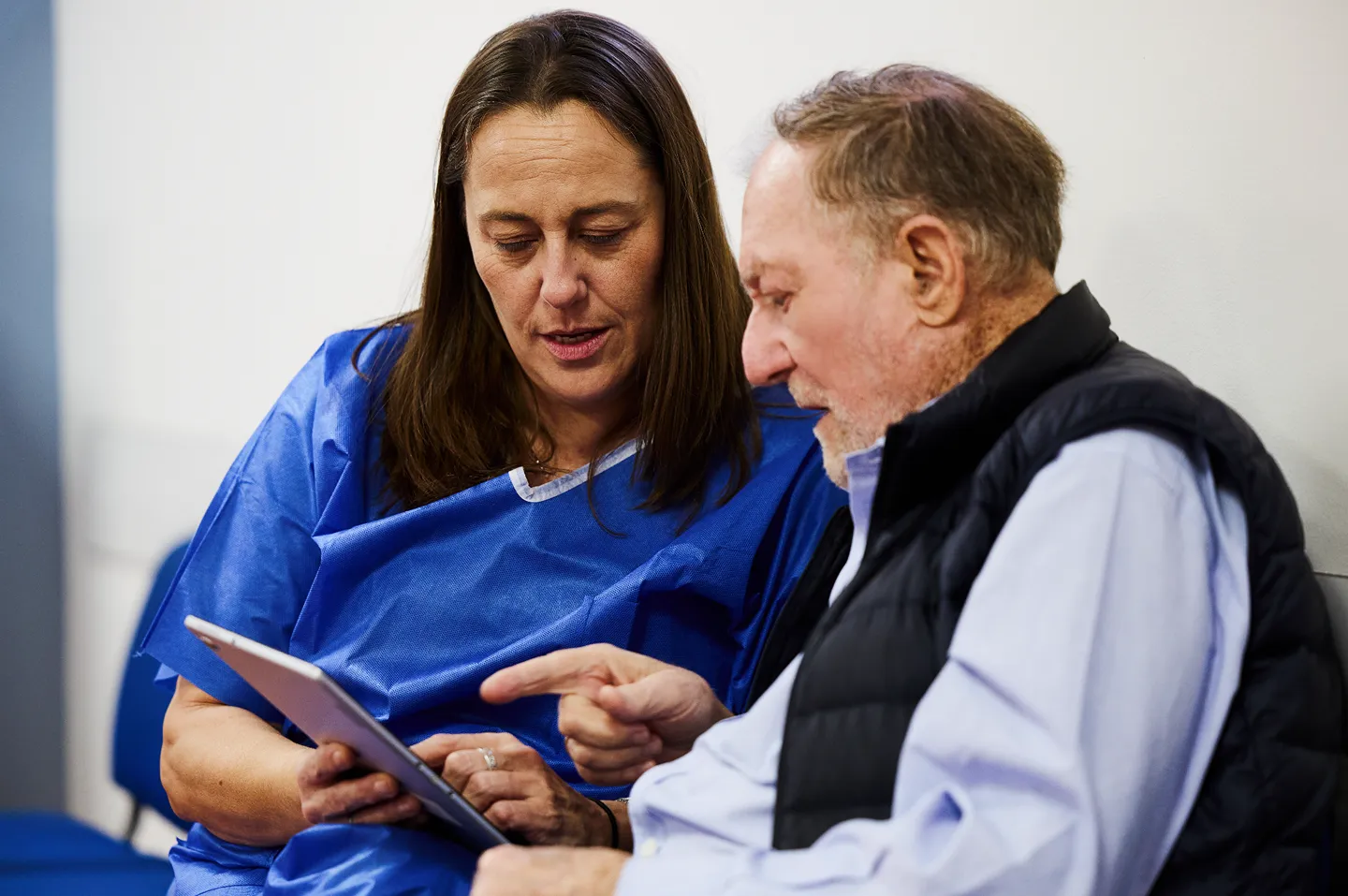 A healthcare worker in protective clothing holds hands and smiles at an older patient receiving medical treatment, while another patient sits nearby in a clinic setting.