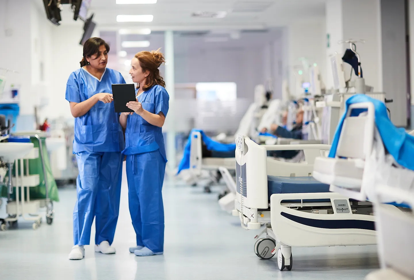 A healthcare worker in protective clothing holds hands and smiles at an older patient receiving medical treatment, while another patient sits nearby in a clinic setting.