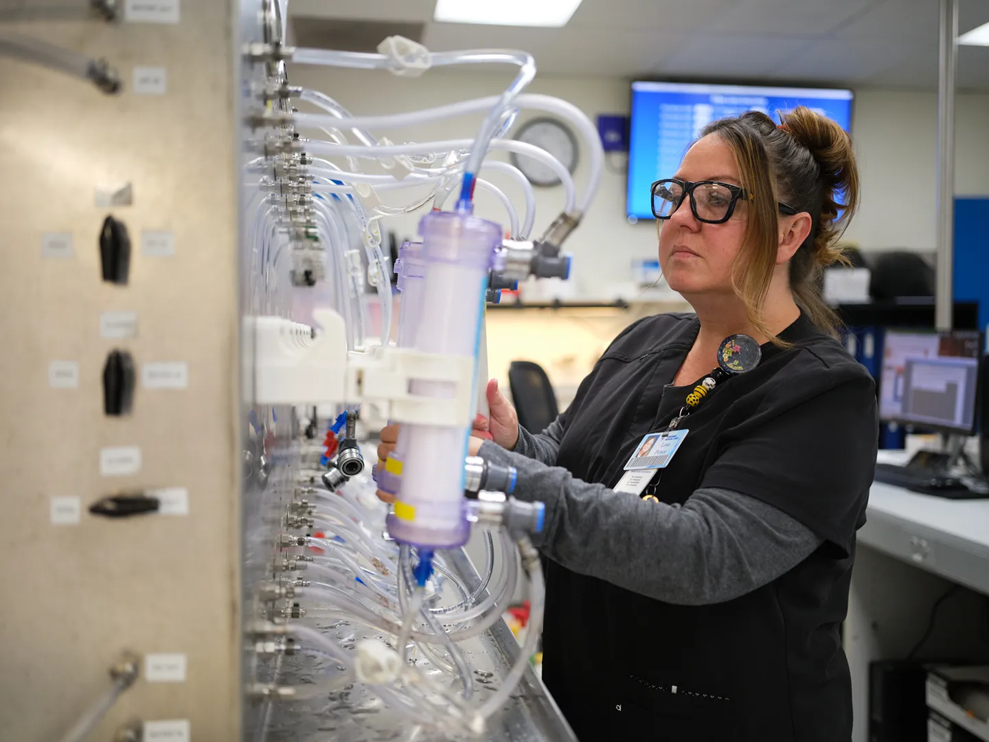 A healthcare worker in protective clothing holds hands and smiles at an older patient receiving medical treatment, while another patient sits nearby in a clinic setting.
