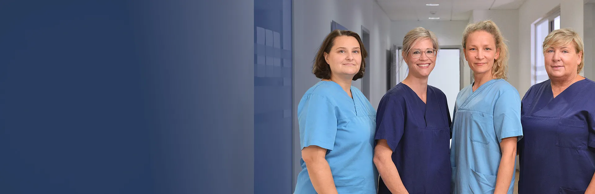Two women in blue medical scrubs smile and look at a tablet together in a healthcare setting, with blurred medical equipment and personnel in the background.