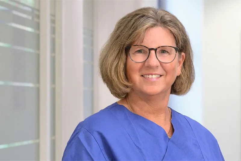 A group of healthcare professionals in blue scrubs stands together in a hospital corridor, smiling at the camera. Some have stethoscopes around their necks, and hospital beds are visible nearby.