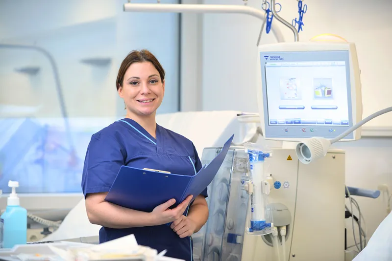 A group of nine healthcare professionals, dressed in blue and white medical scrubs, stand together indoors in front of large windows with a view of greenery outside. They are smiling and posing for the photo.