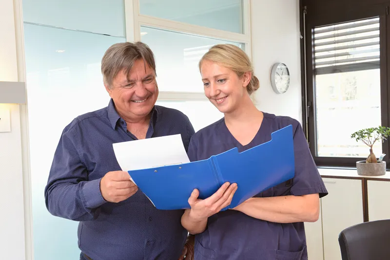 Two men, one in business casual clothes and the other in medical scrubs, smile and bump fists in a bright, modern healthcare setting.