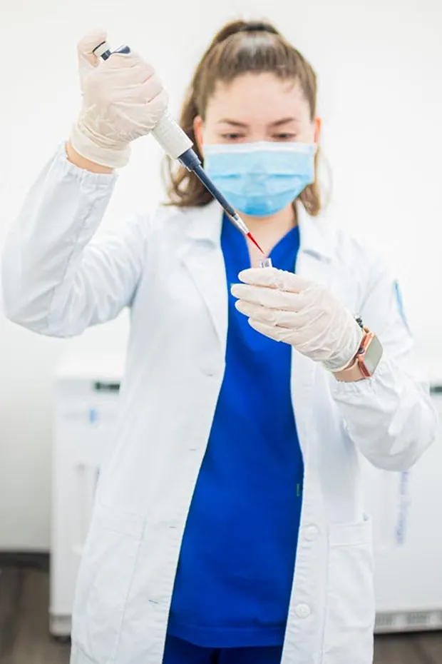 Masked lab associate in a white coat using a pipette to add liquid to a sample vial
