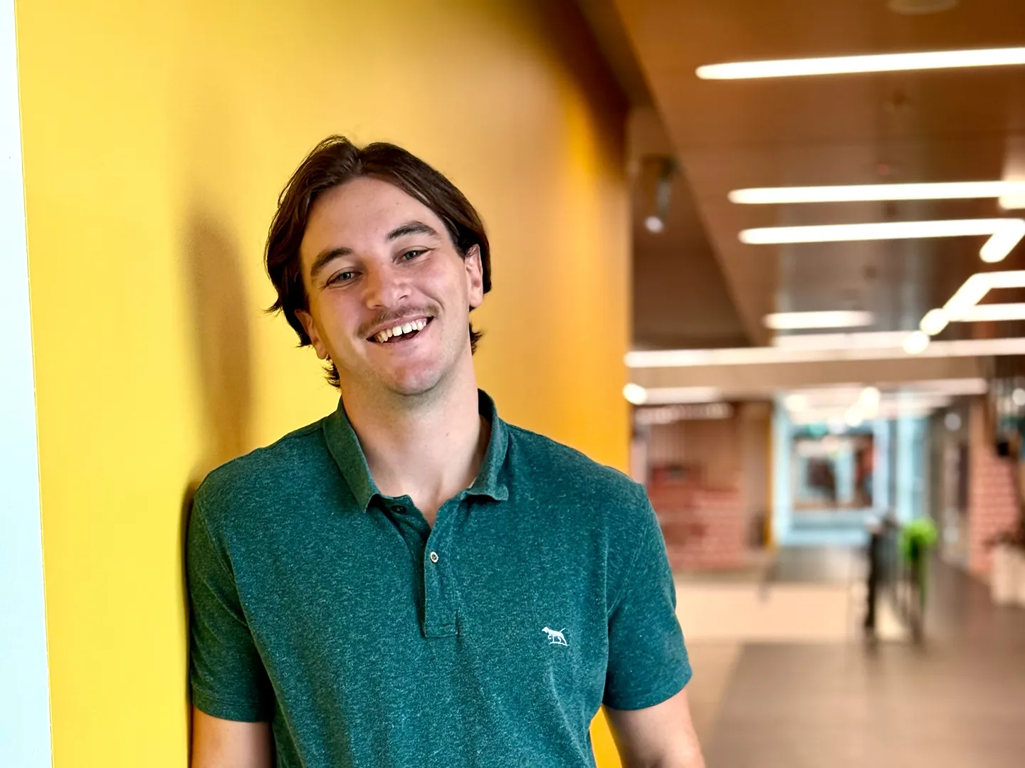 A smiling man in a green shirt, standing beside a yellow wall in an office corridor